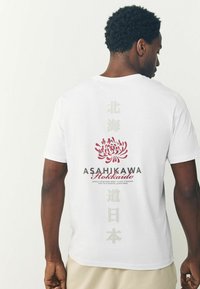 Hvid bomulds T-shirt med et rødt blomstermotiv, teksten "ASAHIKAWA Hokkaido" og lodrette japanske tegn i svag grå.