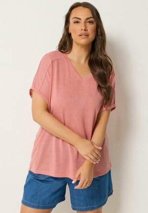 Femme aux longs cheveux bruns portant un haut rose à manches courtes en V et un short en jean bleu, debout devant un fond clair uni.