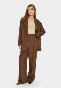 Donna in piedi con una mano in tasca, che indossa un blazer oversize marrone, pantaloni larghi, un top beige e scarpe scure con punta, su sfondo bianco.