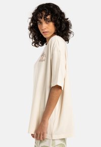 Kremfarget oversized t-skjorte laget av bomull, med korte ermer, rund hals og et trykt design foran. Glatt tekstur.