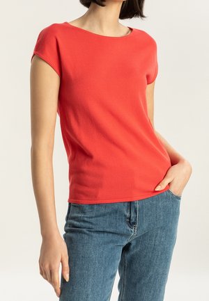 Kobieta w koralowym topie z krótkim rękawem lekko wsuniętym w niebieskie dżinsy, z ręką włożoną do kieszeni na neutralnym tle.