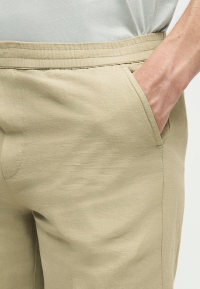 Μπεζ παντελόνι με ελαστική μέση και πλαϊνή τσέπη όπου βγαίνει ένα χέρι, φορεμένο με λευκό πουκάμισο.