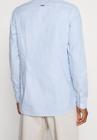 Lichtblauw geweven overhemd met lange mouwen, knoopmanchetten, afgeronde zoom en subtiel ruitpatroon. Heeft een logodetail bij de achterhals.