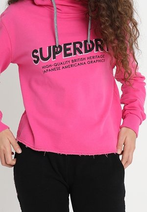 Personne portant un sweat à capuche Superdry rose vif avec des cordons blancs et un pantalon noir, les mains reposant près des poches.