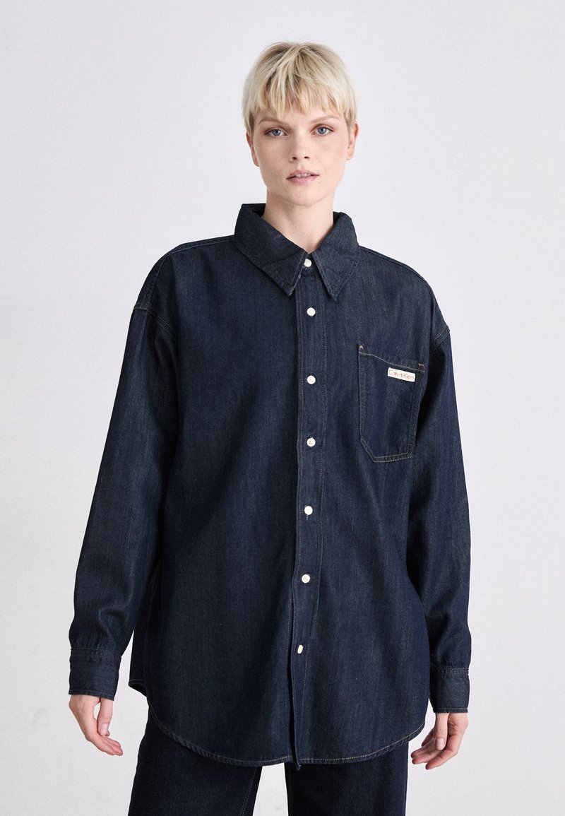 Mørk denim oversized skjorte med knappelukking foran, spiss krage, en brystlomme og lange ermer. Har hvite knapper og søm.