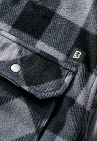Tessuto flanella nero e grigio con un motivo a scacchi. Presenta una chiusura a pressione argentata e un'etichetta nera con un logo bianco. Texture morbida.