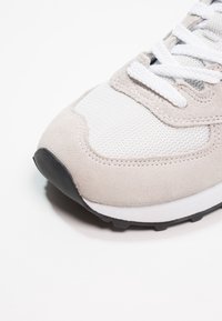 Vaaleanharmaa lenkkari, jossa on teksturoitu mokkanahkapinta, verkkomaiset paneelit, valkoiset nauhat ja musta kumipohja, joka on varustettu pyöreällä kärjellä.