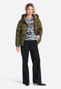 Olijfgroene pufferjack met capuchon, gecombineerd met een multicolored gebreide trui en zwarte wijde broek. Hoge hakken maken de look helemaal af.