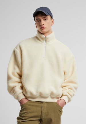 Fleece pullover in cream with a high collar and half-zip, featuring ribbed cuffs. Paired with olive green corduroy pants.