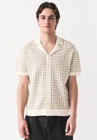 Junger Mann, der ein beige-farbenes, kurzärmeliges Hemd mit geometrischem Quadratmuster und schwarze Hosen trägt, vor einem weißen Hintergrund stehend.