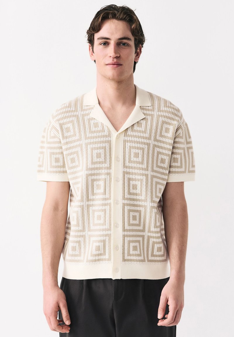 Junger Mann, der ein beige-farbenes, kurzärmeliges Hemd mit geometrischem Quadratmuster und schwarze Hosen trägt, vor einem weißen Hintergrund stehend.