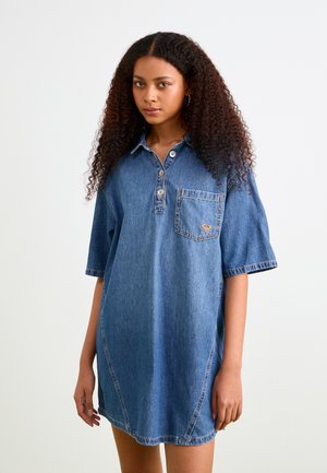 Denimjurk met korte mouwen, kraagontwerp, voorzak en oranje stikdetails. Middelblauwe kleur en een relaxte pasvorm.