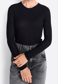 Schwarzes Langarmoberteil mit gerippter Textur und Rundhalsausschnitt. Kombiniert mit hochgeschnittenen grauen Jeans, Hände auf dem Bund abstützend.
