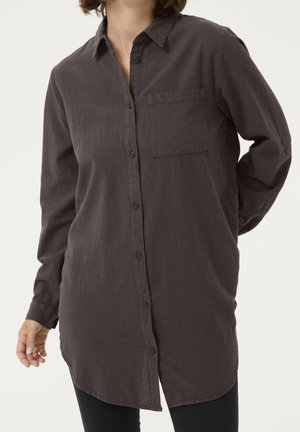 Personne portant une longue chemise boutonnée gris foncé avec une poche poitrine, debout devant un fond clair uni avec une main derrière le dos.