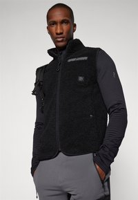 Μαύρο γιλέκο φλις με φερμουάρ μπροστά, ψηλό γιακά, υφασμάτινη επιφάνεια και μπροστινή τσέπη. Φορεμένο πάνω από μια μαύρη μπλούζα με μακριά μανίκια.