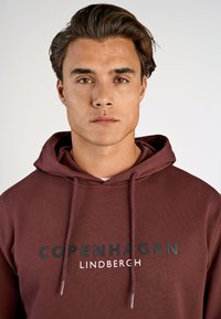 Hoodie color bordeaux con tasca frontale, cappuccio con cordino e testo stampato "COPENHAGEN LINDBERCH" in contrasto tra nero e bianco.