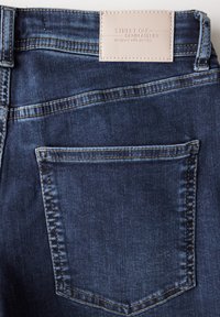 Tmavě modré džíny mají koženou etiketu se zašitou textem "STREET ONE DENIM ATELIER" na pase a jedno zadní kapsu.