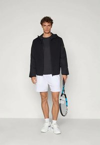 Jaunas vyras stovi, vilkėdamas juodą zip-up švarkinę, juodą marškinėlį, baltus šortus, baltas sportbačius, laikydamas teniso raketę šone prieš paprastą fono.