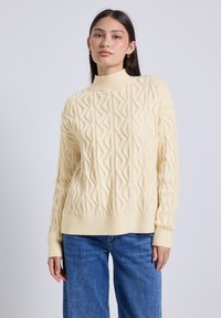 Jeune femme portant un pull col roulé en tricot torsadé crème et un jean bleu, se tenant contre un fond gris clair uni.
