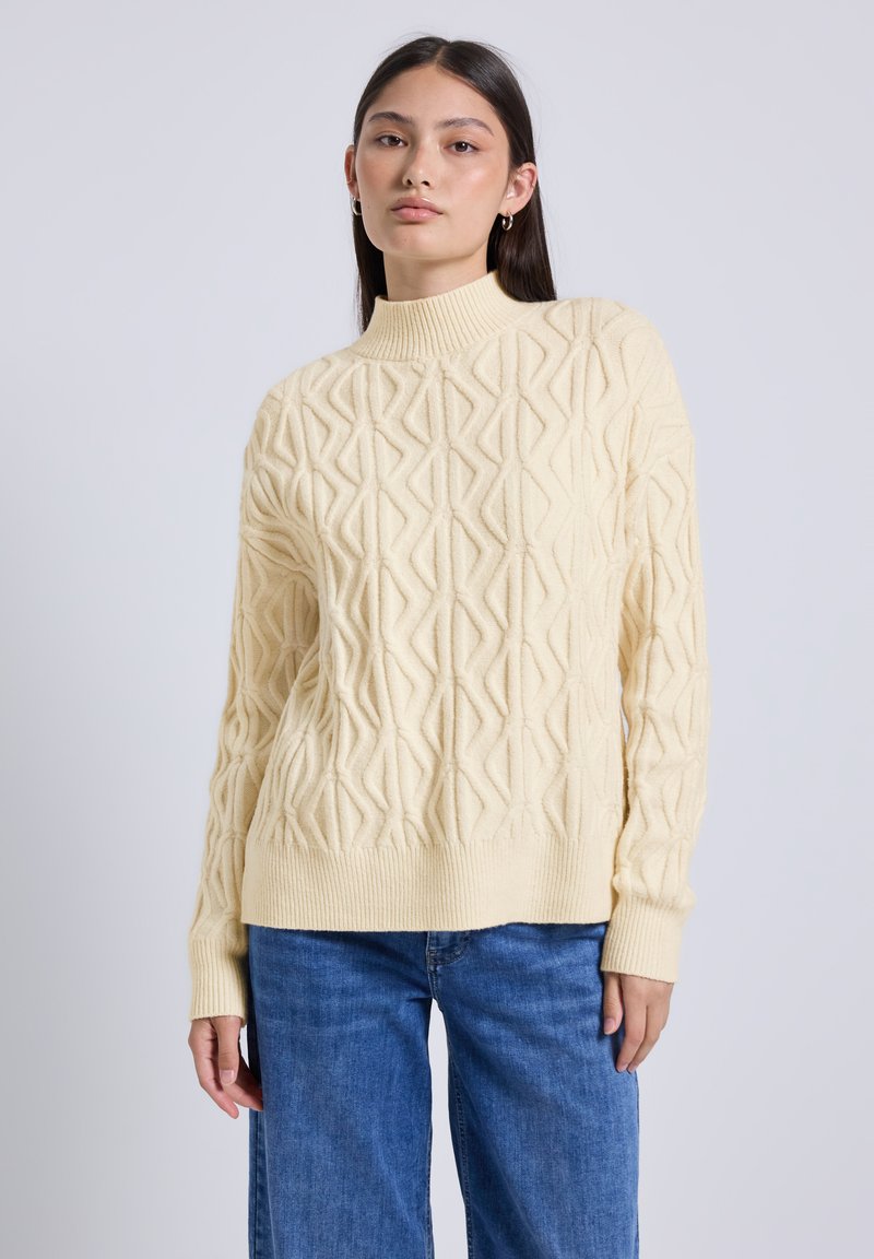 Jeune femme portant un pull col roulé en tricot torsadé crème et un jean bleu, se tenant contre un fond gris clair uni.