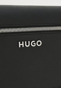 Portafoglio in pelle nera con chiusura zip in argento, logo "HUGO" in rilievo e superficie testurizzata con motivi sottili.