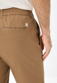 Pantalon en tissu marron avec une ceinture élastique, comportant une poche arrière avec un écusson logo carré et une surface lisse et texturée.