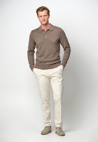 Brauner Strickpullover mit Polokragen, beigefarbene Hose und hellbeige Sneaker. Das Model steht mit den Händen in den Taschen vor einem weißen Hintergrund.