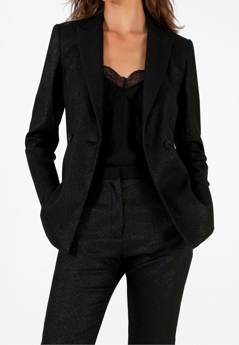 Femme portant un blazer noir pailleté ajusté et un pantalon assorti sur un caraco noir à dentelle, les mains dans les poches.