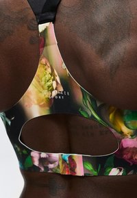 Κοντινή λήψη σκοτεινής επιδερμίδας στην πλάτη που φορά φλοράλ αθλητικό σουτιέν με άνοιγμα-κλειδί και λογότυπο Ted Baker Sport.