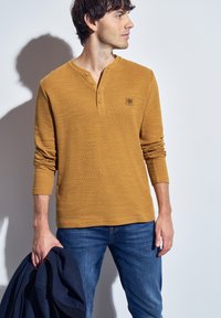 Muž oblečený v hořčicově žlutém dlouhém rukávu henley tričku a modrých džínách, držící námořnickou bundu v levé ruce, dívající se doprava.