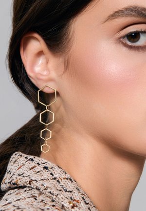 Złote geometryczne kolczyki z interlinked hexagonalnymi kształtami, o gładkim wykończeniu i długim, wiszącym fasonie. Widoczny teksturowany kołnierz.