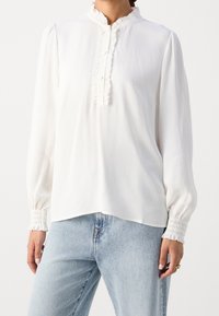 Kvinde iført en hvid bluse med lange ærmer og flæser ved kraven og manchetterne, parret med lyseblå jeans, der viser overkrop og hænder.