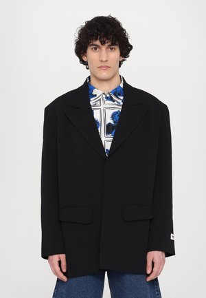 Schwarzer Blazer mit oversized Passform, strukturierten Schultern und Reverskragen. Verfügt über zwei Fronttaschen und eine glatte Stofftextur.