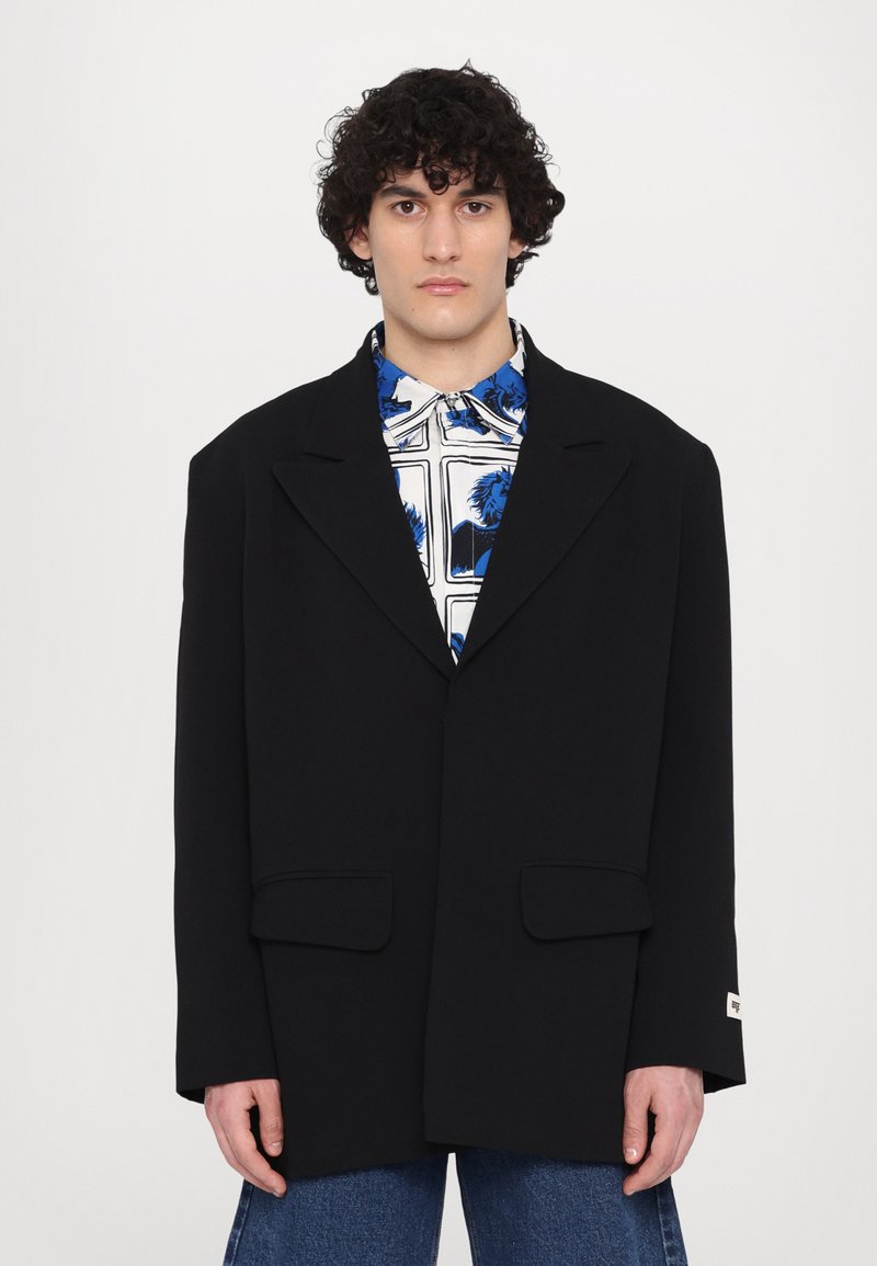 Black blazer with oversized fit, structured shoulders, and notch lapels. Features two front pockets and a smooth fabric texture.