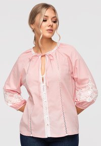 Femme portant un chemisier rose clair avec des détails en dentelle blanche, manches trois-quarts et un lien au niveau du col, posant avec les mains sur les hanches.