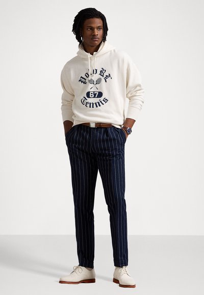 Creme hoodie med blå grafisk print, marineblå stribede bukser og lyse beige sko. Outfittet har en afslappet pasform og kontrasterende farver.