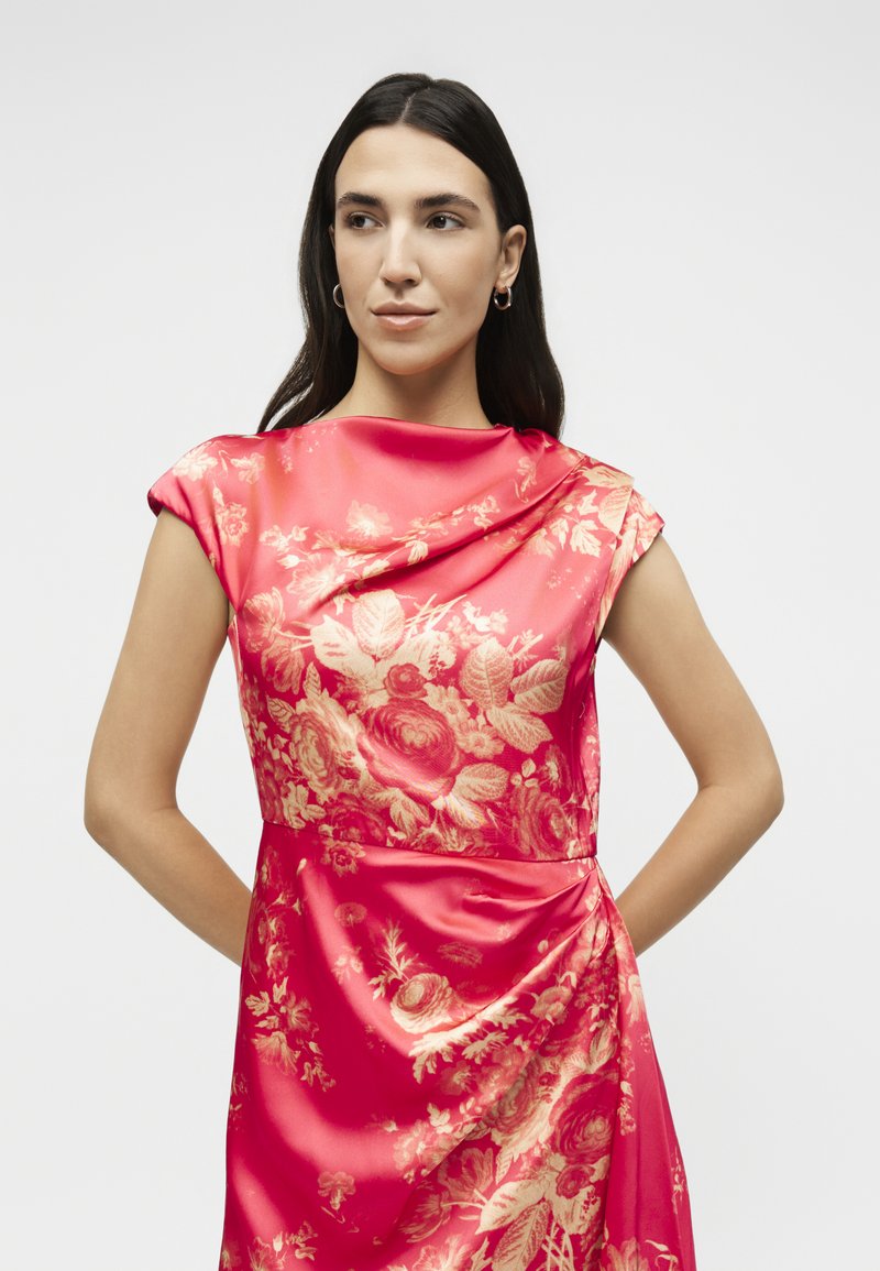 Donna con capelli lunghi e scuri, con orecchini a cerchio e un vestito di raso rosso con motivi floreali dorati, in piedi con le mani dietro la schiena.