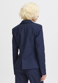 Námořnický modrý blazer s přiléhavým střihem. Má strukturovaná ramena, klopový límec a manžety na tři knoflíky. Hladká tkanina s čistým designem.