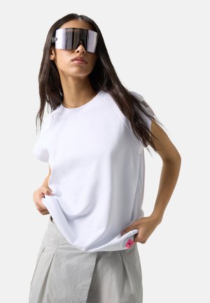 Kobieta nosząca duże lustrzane okulary przeciwsłoneczne, białą koszulę z krótkim rękawem i jasnoszarą plisowaną spódnicę, trzymająca brzeg koszuli.