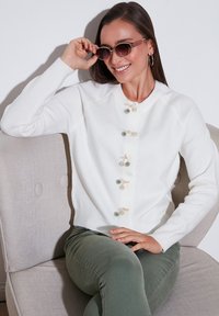 Cárdigan blanco de punto con botones decorativos que presentan pompones en crema y verde. La modelo lleva gafas de sol, combinadas con pantalones verde oliva.