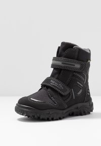 Botas negras hasta el tobillo con un exterior de tejido texturizado, correa de Velcro y suela de goma con patrones de relieve detallados para tracción.