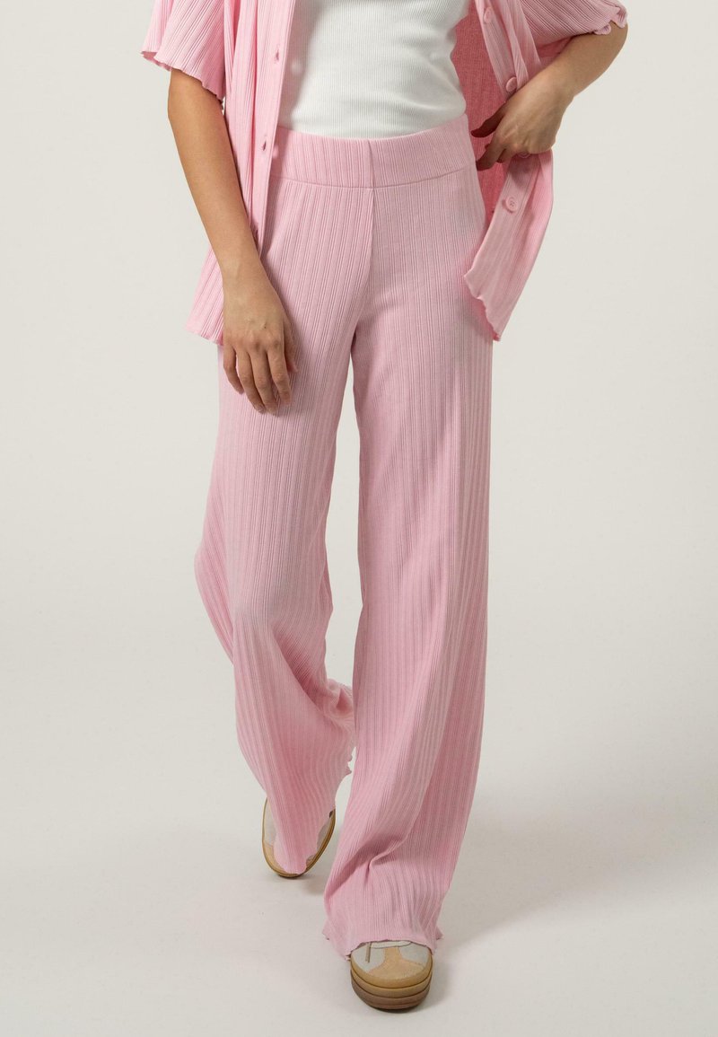 Pantalons larges rose clair avec une texture côtelée verticale, taille haute et une coupe fluide. Associés à un haut blanc et des chaussures décontractées.