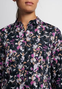 Camisa de botón con un fondo negro y un patrón floral en tonos de rosa, blanco y beige. Diseño con cuello y botones.