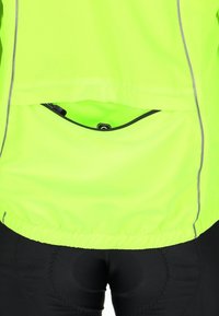 Fluorescerende gul sportsjakke med refleks detaljer, elastisk kant og en lynlåslomme på ryggen. Kombination med sorte leggings.