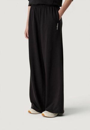 Personne portant un pantalon noir à jambes larges avec une taille élastique et des baskets blanches avec des semelles beiges, debout avec une main dans la poche.
