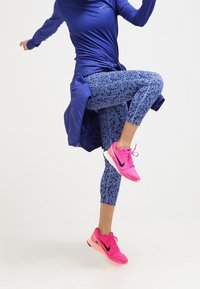 Niebieska sportowa bluzka z długim rękawem i węzłem, wzorzyste niebieskie legginsy oraz intensywnie różowe sneakersy z czarnymi akcentami.