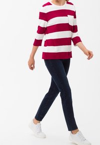 Top de manga larga a rayas rojas y blancas con un bolsillo frontal, combinado con jeans azul oscuro y zapatillas blancas. Tejido ligero, ajuste casual.
