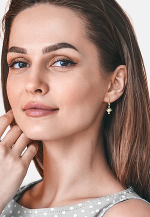 Goldene Anhängerohrringe mit geometrischem Design und einem kleinen Steinelement. Ein Modell mit langen, glatten braunen Haaren trägt ein gepunktetes Oberteil.