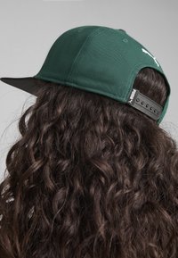 Căciulă verde cu vizieră neagră, curea ajustabilă în spate, având un logo pe lateral. Materialul pare texturat, cu coaseri vizibile.