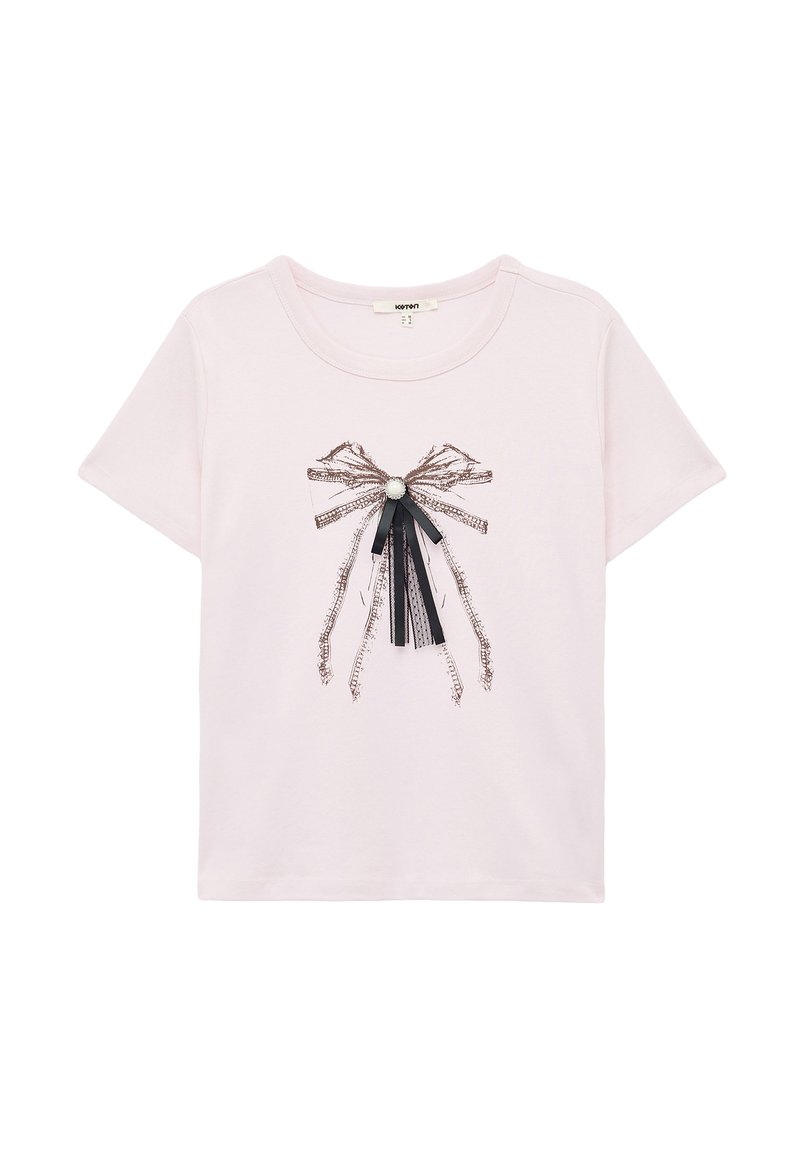 Camiseta de manga corta color rosa claro con un lazo decorativo de cintas negras y plateadas y una gema blanca central. Tejido suave, corte casual.